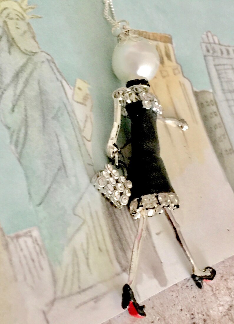 May include: A silver necklace with a pearl and a black and silver figurine of a woman wearing a dress and carrying a purse. The figurine is adorned with rhinestones.