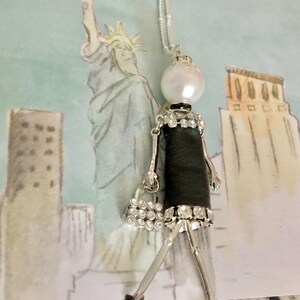 May include: A silver chain necklace with a black and white doll pendant. The doll is wearing a black dress with rhinestones and a small purse. The doll has a pearl head and red shoes.