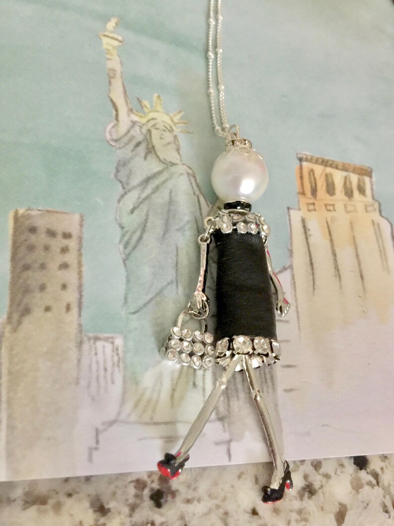 May include: A silver chain necklace with a black and white doll pendant. The doll is wearing a black dress with rhinestones and a small purse. The doll has a pearl head and red shoes.