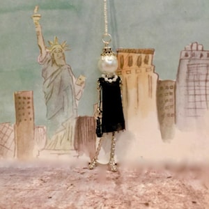 Puede incluir: Un collar de plata con un vestido negro y un dije de perla. El dije es una pequeña figura estilizada de una mujer con un vestido negro. El collar está colgado sobre un fondo de acuarela de un horizonte de ciudad.