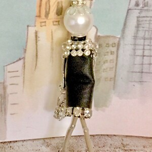 May include: A silver necklace with a black and white figurine pendant. The figurine is wearing a black dress with a rhinestone trim and red high heels. The figurine has a pearl head and a silver chain.