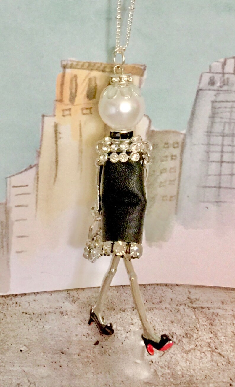 May include: A silver necklace with a black and white figurine pendant. The figurine is wearing a black dress with a rhinestone trim and red high heels. The figurine has a pearl head and a silver chain.