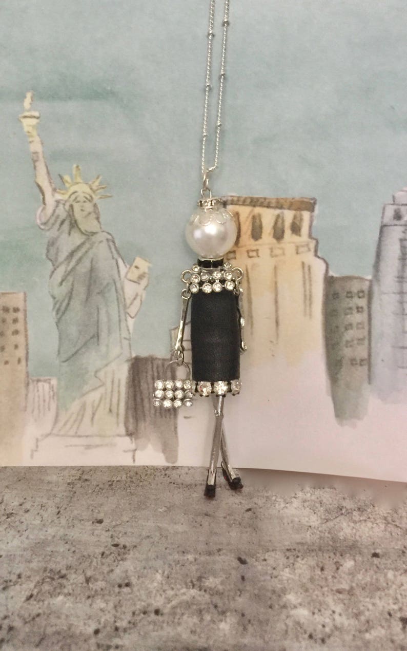 May include: A silver chain necklace with a black and white figurine pendant. The figurine is wearing a black dress with rhinestones and holding a small purse with rhinestones. The pendant is attached to a pearl.