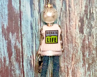 May include: A silver chain necklace with a charm of a woman wearing a white shirt with the words "Donate Life" on it. The woman is wearing blue jeans and has a pearl head.