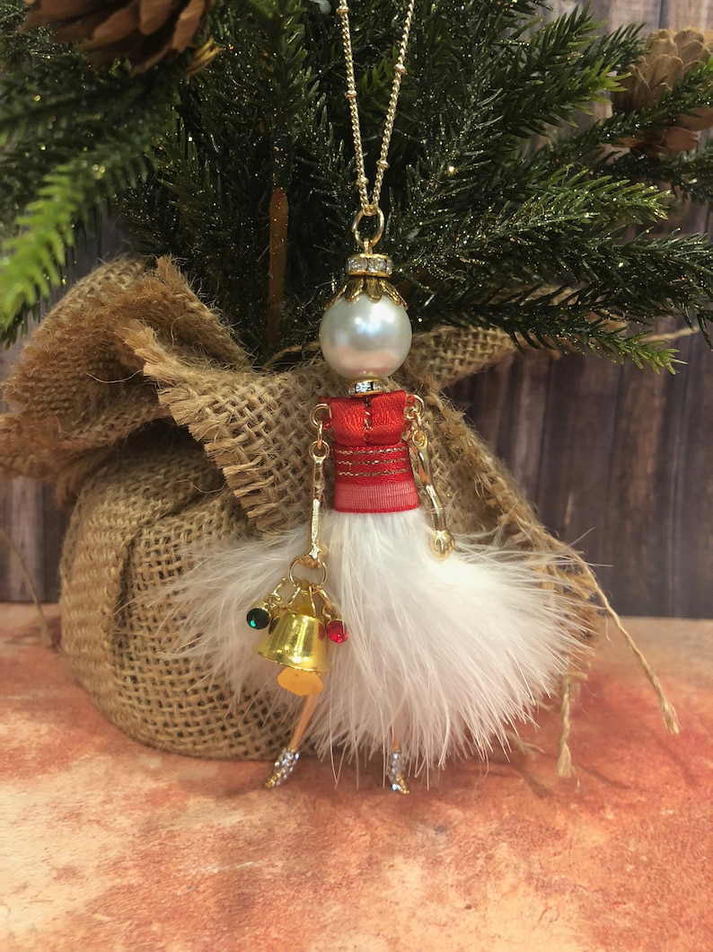 May include: A white feather Christmas ornament with a red and gold dress, a pearl head, and gold bells.