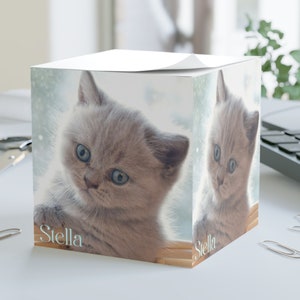 May include: A white cube-shaped notepad with a grey kitten on the front. The kitten has blue eyes and is looking at the camera. The notepad has the word "Stella" printed on the front.