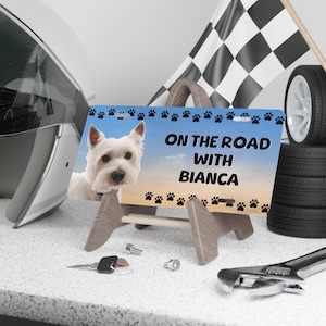 Puede incluir: Un letrero decorativo con un perro blanco y el texto "ON THE ROAD WITH BIANCA" sobre un fondo azul y amarillo. El letrero está sostenido por un caballete de madera, con huellas de patas alrededor del borde.