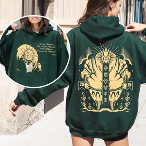 May include: Dark green hoodie with a large graphic on the back featuring a stylized figure with a sunburst design and the numbers 20 and 25. The front has a smaller graphic and text that reads "I too am 'Whooo.' But I'm also 'Wheee!' So the 'Wheee' balances the 'Whooo.' -Esquie."