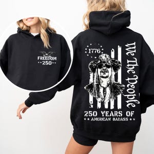 May include: Black hooded sweatshirt with a graphic on the front that says "FREEDOM **250**" and a graphic of an eagle. The back of the sweatshirt has a portrait and the text "We The People" and "250 YEARS OF ** AMERICAN BADASS**".