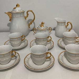 May include: A white porcelain tea set with gold accents. The set includes a coffee pot, sugar bowl, creamer, cups, and saucers. The cups and saucers have a gold border and a gold grid pattern. The handles on the cups, pot, and creamer are gold.