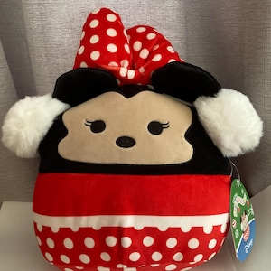 Op de afbeelding: Een Minnie Mouse knuffel met een rode strik met witte stippen, zwarte oren en een rode en witte stippen body. Het speelgoed heeft een witte, donzige rand en een productlabel.