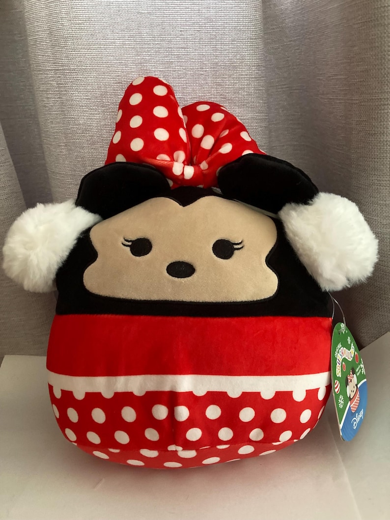 Op de afbeelding: Een Minnie Mouse knuffel met een rode strik met witte stippen, zwarte oren en een rode en witte stippen body. Het speelgoed heeft een witte, donzige rand en een productlabel.