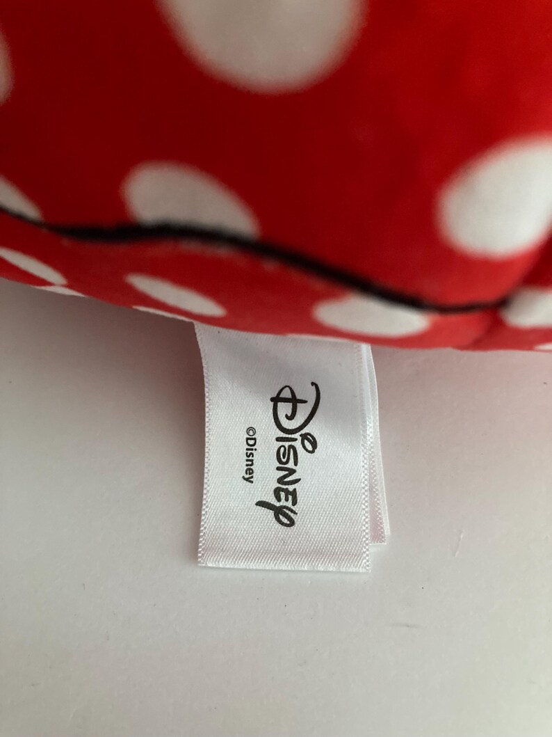 Op de afbeelding: Een close-up van rode stof met witte stippen. Een wit label met het Disney-logo en het woord "Disney" is aan de stof bevestigd. Het label bevat ook het copyrightsymbool.