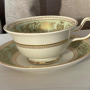 Könnte beinhalten: Ein weißes Tassen- und Untertassen-Set mit goldenen Verzierungen und einem hellgrünen Blumendesign. Die Tasse hat einen gebogenen Henkel und steht auf einer passenden Untertasse. Das Set ist wahrscheinlich aus Porzellan oder feinem Porzellan.