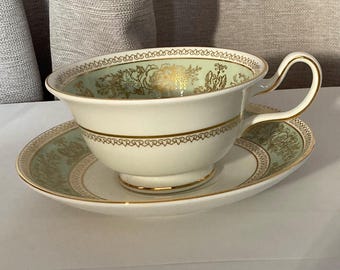 Può includere: Un set di tazza e piattino bianchi con finiture dorate e un motivo floreale verde chiaro. La tazza ha un manico curvo e poggia su un piattino abbinato. Il set è probabilmente in porcellana o porcellana fine.
