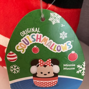 Op de afbeelding: Een groen label met de tekst "Original Squishmallows" en een Minnie Mouse-afbeelding. Het label heeft sneeuwvlok- en kerstbalontwerpen. Het Disney-logo staat onderaan. Het pluche speelgoed is rood, zwart en wit.