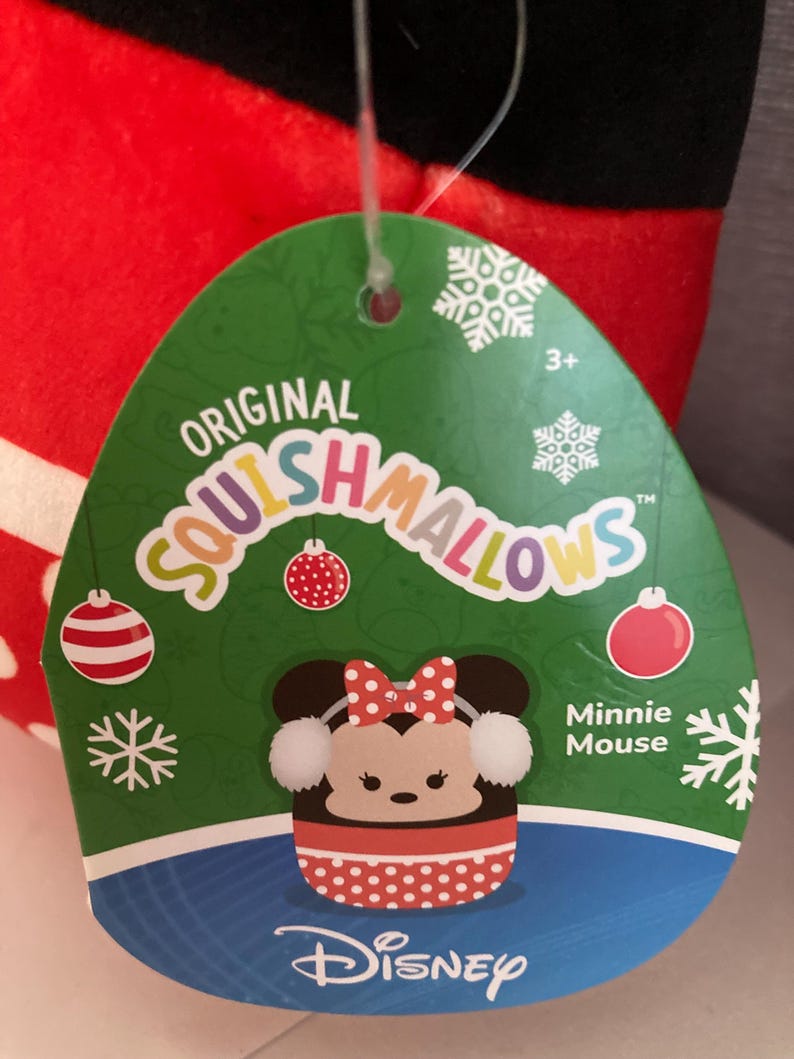 Op de afbeelding: Een groen label met de tekst "Original Squishmallows" en een Minnie Mouse-afbeelding. Het label heeft sneeuwvlok- en kerstbalontwerpen. Het Disney-logo staat onderaan. Het pluche speelgoed is rood, zwart en wit.
