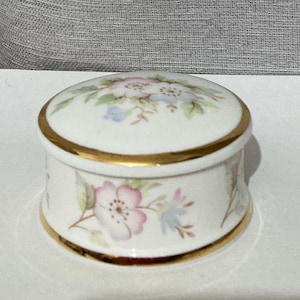 May include: A small, round, white porcelain trinket box with a lid. The box is decorated with pink and blue floral designs and gold trim. The lid also features a floral pattern. The box is likely used for storing small jewellery items.