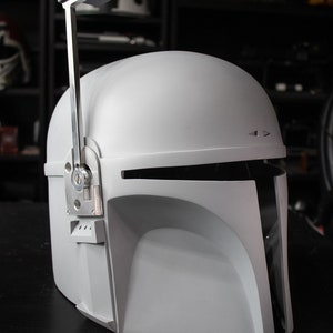 May include: A light gray Mandalorian helmet with a visor and a silver-colored antenna. The helmet has a rounded top and a distinctive T-shaped visor. The antenna is attached to the top of the helmet with a hinge.
