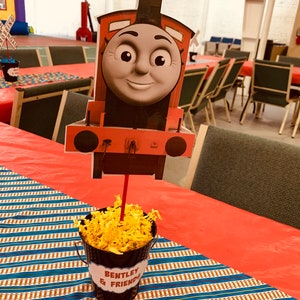 May include: Red and black cartoon train engine centerpiece with a smiling face. The train is on a red and blue striped table runner with a black bucket filled with yellow shredded paper. The bucket has a white label that says "Bentley & Friends".