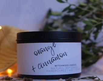 May include: A black candle tin with a white label that reads "orange + cinnamon". The label also includes the text "Witchwood Candles", "Natural Soy Candle", and "Handpoured | 6oz | 180g".