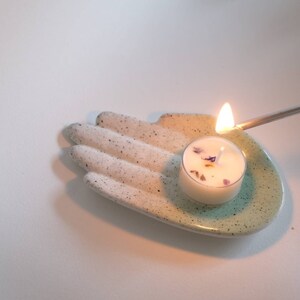 Puede incluir: Un plato de cer&aacute;mica blanco con forma de mano con un glaseado turquesa en la palma. Una peque&ntilde;a vela de t&eacute; blanca con flores secas en su interior est&aacute; sentada en la palma de la mano y se est&aacute; encendiendo con una cerilla.