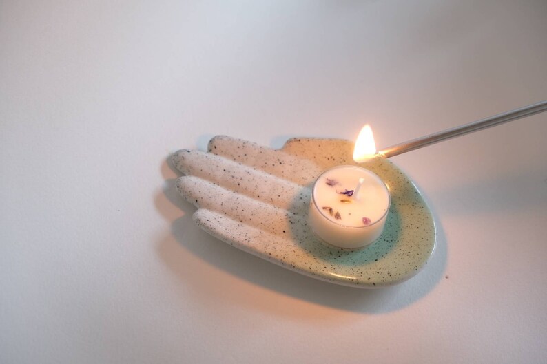 Puede incluir: Un plato de cer&aacute;mica blanco con forma de mano con un glaseado turquesa en la palma. Una peque&ntilde;a vela de t&eacute; blanca con flores secas en su interior est&aacute; sentada en la palma de la mano y se est&aacute; encendiendo con una cerilla.