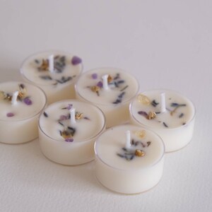 Puede incluir: Seis velas de t&eacute; blancas con flores y hierbas secas incrustadas en la cera. Las velas est&aacute;n dispuestas en c&iacute;rculo sobre una superficie blanca.