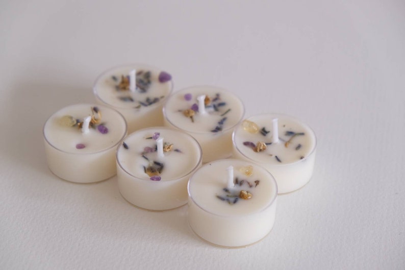 Puede incluir: Seis velas de t&eacute; blancas con flores y hierbas secas incrustadas en la cera. Las velas est&aacute;n dispuestas en c&iacute;rculo sobre una superficie blanca.
