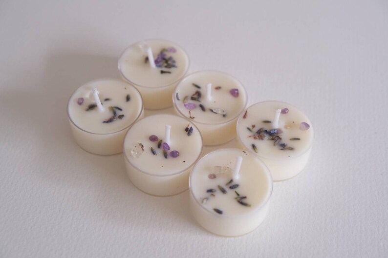 Puede incluir: Seis velas de t&eacute; blancas con lavanda y cristales morados incrustados en la cera. Las velas est&aacute;n dispuestas en c&iacute;rculo sobre una superficie blanca.