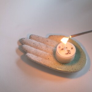 Puede incluir: Un plato de cer&aacute;mica blanco con forma de mano y un esmalte verde azulado contiene una peque&ntilde;a vela de t&eacute; blanca. Un encendedor plateado largo se utiliza para encender la vela.
