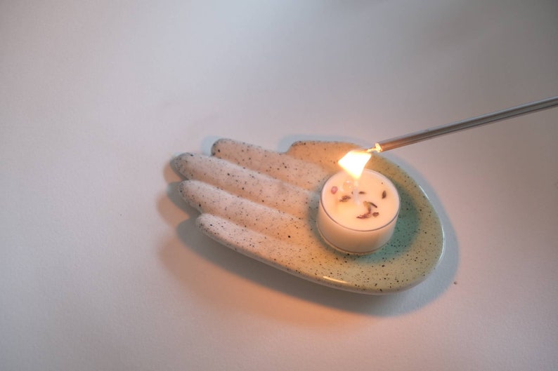 Puede incluir: Un plato de cer&aacute;mica blanco con forma de mano y un esmalte verde azulado contiene una peque&ntilde;a vela de t&eacute; blanca. Un encendedor plateado largo se utiliza para encender la vela.