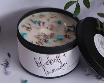 May include: A round, black candle tin with a white label that reads "bluebell + lavender". The candle is filled with white wax and decorated with blue, purple, and turquoise stones and dried flowers. The lid is open.