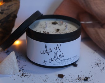 May include: A black candle jar with a white label that reads "wake up + coffee". The candle is surrounded by coffee beans and a white stone.