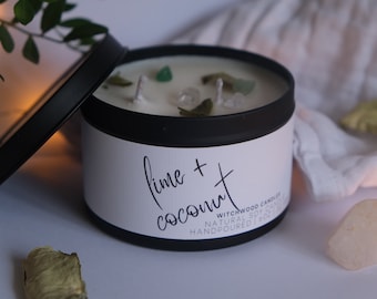 May include: A black candle jar with a white label that reads "lime + coconut" and "Witchwood Candles" in a handwritten font. The candle is made with natural soy wax and is hand-poured. The jar is partially open, revealing a white candle with green and white crystals embedded in the wax.