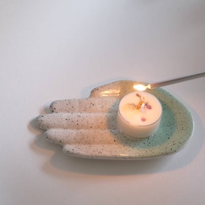 Puede incluir: Un plato de cer&aacute;mica blanco con forma de mano con un glaseado turquesa en el borde. Una peque&ntilde;a vela de t&eacute; blanca con flores secas en su interior se est&aacute; encendiendo con un encendedor plateado largo.