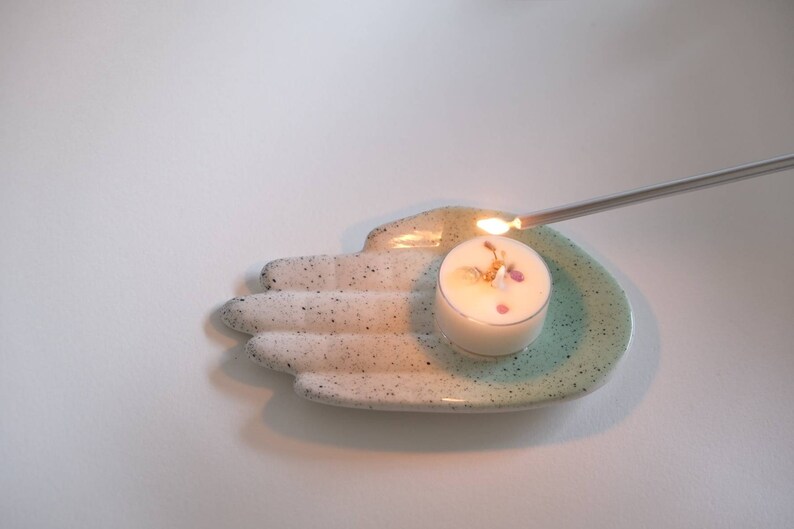 Puede incluir: Un plato de cer&aacute;mica blanco con forma de mano con un glaseado turquesa en el borde. Una peque&ntilde;a vela de t&eacute; blanca con flores secas en su interior se est&aacute; encendiendo con un encendedor plateado largo.
