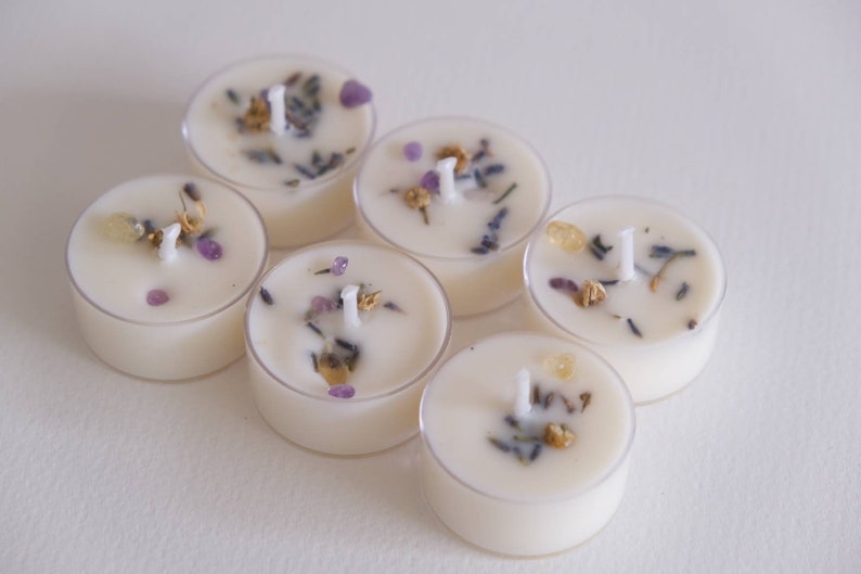 Puede incluir: Seis peque&ntilde;as velas blancas para t&eacute; con flores secas y peque&ntilde;as piedras moradas y doradas incrustadas. Las velas est&aacute;n dispuestas en c&iacute;rculo sobre una superficie blanca.