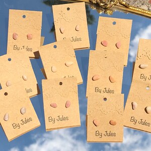 May include: A collection of small, natural stone earrings displayed on brown cards. Each card has the text "By Jules" printed on it. The earrings are in various shades of pink and beige.