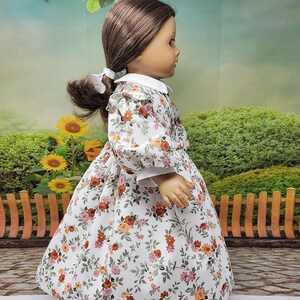 K&ouml;nnte beinhalten: Puppe in einem wei&szlig;en Kleid mit einem Blumenmuster aus orangefarbenen, rosa und gr&uuml;nen Blumen. Das Kleid hat einen wei&szlig;en Kragen und Manschetten. Die braunen Haare der Puppe sind zu einem Pferdeschwanz mit einer wei&szlig;en Schleife gebunden.