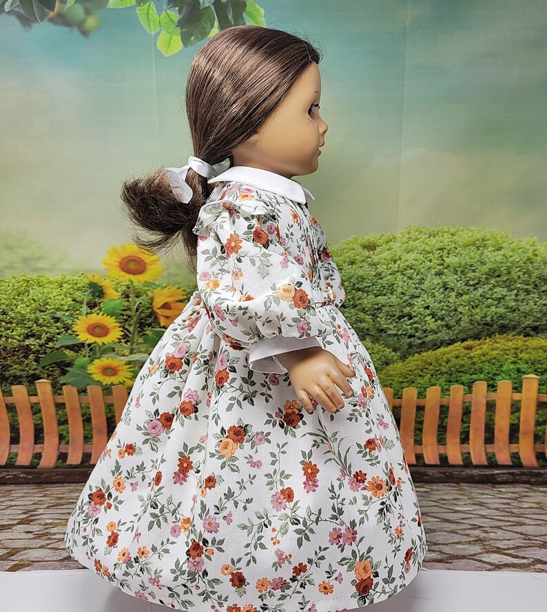 K&ouml;nnte beinhalten: Puppe in einem wei&szlig;en Kleid mit einem Blumenmuster aus orangefarbenen, rosa und gr&uuml;nen Blumen. Das Kleid hat einen wei&szlig;en Kragen und Manschetten. Die braunen Haare der Puppe sind zu einem Pferdeschwanz mit einer wei&szlig;en Schleife gebunden.