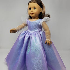 May include: A doll wearing a lavender princess gown with a shimmering effect. The dress has a full skirt and a fitted bodice. The doll also wears a silver crown and has brown hair.