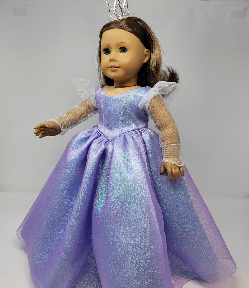 May include: A doll wearing a lavender princess gown with a shimmering effect. The dress has a full skirt and a fitted bodice. The doll also wears a silver crown and has brown hair.