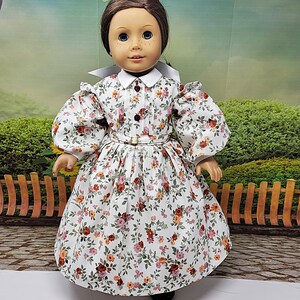 K&ouml;nnte beinhalten: Puppe in einem wei&szlig;en Kleid mit Blumenmuster, wei&szlig;em Kragen und Puff&auml;rmeln. Das Kleid hat ein eng anliegendes Oberteil, eine Taille mit G&uuml;rtel und einen weiten Rock. Die Puppe hat braune Haare und blaue Augen. Der Hintergrund ist eine verschwommene Szene im Freien.