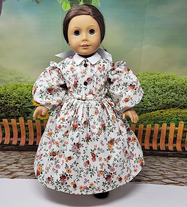 K&ouml;nnte beinhalten: Puppe in einem wei&szlig;en Kleid mit Blumenmuster, wei&szlig;em Kragen und Puff&auml;rmeln. Das Kleid hat ein eng anliegendes Oberteil, eine Taille mit G&uuml;rtel und einen weiten Rock. Die Puppe hat braune Haare und blaue Augen. Der Hintergrund ist eine verschwommene Szene im Freien.