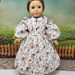 K&ouml;nnte beinhalten: Eine Puppe in einem wei&szlig;en Kleid mit floralem Muster, wei&szlig;em Kragen und Puff&auml;rmeln. Das Kleid hat ein eng anliegendes Oberteil, eine Taille mit G&uuml;rtel und einen weiten Rock. Das Blumenmuster umfasst rote, orangefarbene und rosa Blumen.