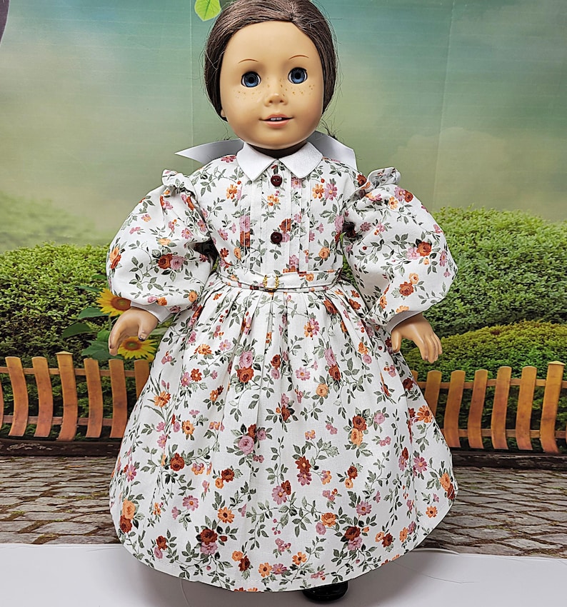 K&ouml;nnte beinhalten: Eine Puppe in einem wei&szlig;en Kleid mit floralem Muster, wei&szlig;em Kragen und Puff&auml;rmeln. Das Kleid hat ein eng anliegendes Oberteil, eine Taille mit G&uuml;rtel und einen weiten Rock. Das Blumenmuster umfasst rote, orangefarbene und rosa Blumen.