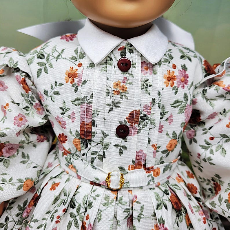 K&ouml;nnte beinhalten: Puppe in einem wei&szlig;en Kleid mit floralem Muster, wei&szlig;em Kragen und Puff&auml;rmeln. Das Kleid hat einen kleinen G&uuml;rtel mit goldener Schnalle und braunen Kn&ouml;pfen. Das Blumenmuster enth&auml;lt rosa, orange und gr&uuml;ne Akzente.