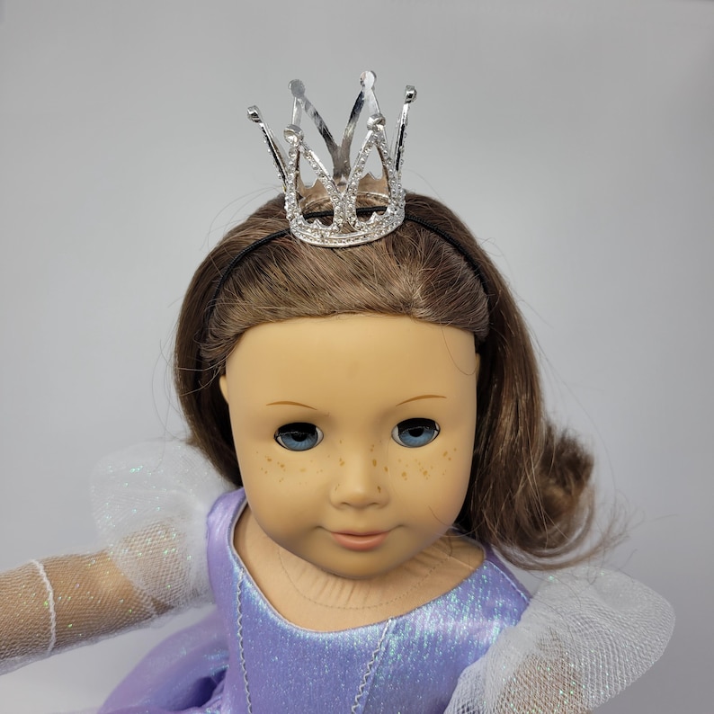 May include: A doll with brown hair, blue eyes, and freckles, wearing a silver crown and a lavender dress with white sleeves. The crown is attached to a black headband.