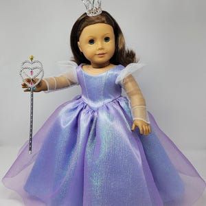 May include: A doll dressed as a princess, wearing a shimmering lavender gown with a sheer overlay. The doll has brown hair, blue eyes, and a silver crown. She holds a silver wand with a heart-shaped top, perfect for imaginative play.